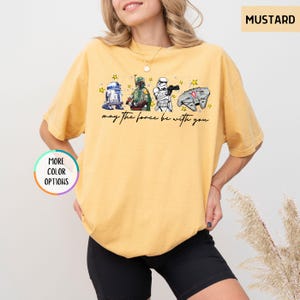 Puede incluir: Camiseta color mostaza con un gr&aacute;fico de Star Wars. El dise&ntilde;o incluye a R2-D2, Boba Fett, un Stormtrooper y el Halc&oacute;n Milenario, con el texto "may the force be with you".