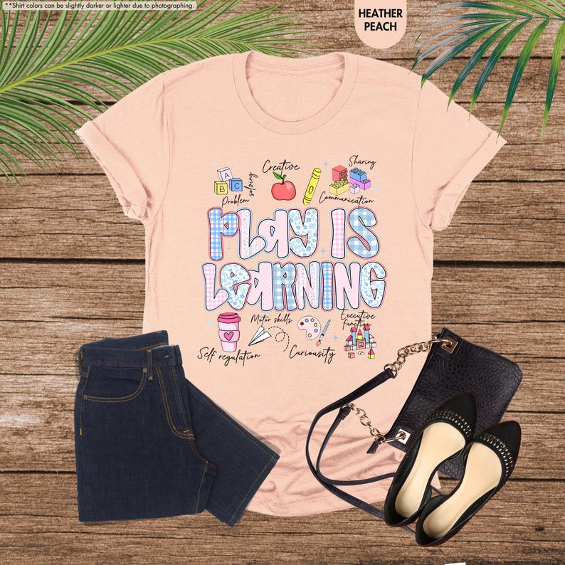 May include: A peach-coloured t-shirt with the words "PLAY IS LEARNING" in a colourful design. The image includes dark blue jeans, black flats, and a black handbag. The shirt features educational graphics like an apple, pencil, and building blocks.