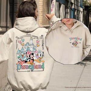 May include: A cream-colored hoodie with a graphic of Mickey Mouse and friends in front of Disneyland. The text "Disneyland Est. 1955" is printed below the graphic. The hoodie has a drawstring hood and a kangaroo pocket.