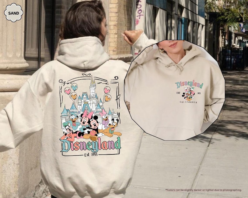 May include: A cream-colored hoodie with a graphic of Mickey Mouse and friends in front of Disneyland. The text "Disneyland Est. 1955" is printed below the graphic. The hoodie has a drawstring hood and a kangaroo pocket.
