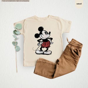 May include: A cream-colored t-shirt featuring a classic Mickey Mouse graphic in black, red, and white. Paired with brown pants. The image has a natural aesthetic.