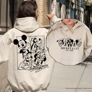 Pu&ograve; includere: Felpa con cappuccio color sabbia con grafica in bianco e nero di Topolino e amici. Il retro mostra Topolino, Pippo e Minnie, mentre il davanti presenta la scritta "Mickey & Co. Est. 1928."