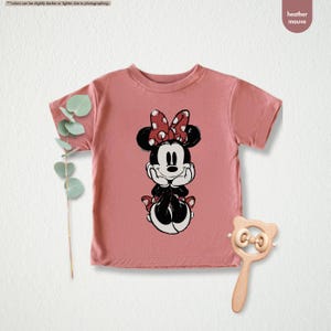 May include: Heather mauve t-shirt featuring a Minnie Mouse graphic. The graphic shows Minnie Mouse in black and white with a red polka dot bow. The shirt is short-sleeved and has a round neckline. A wooden rattle is next to the shirt.
