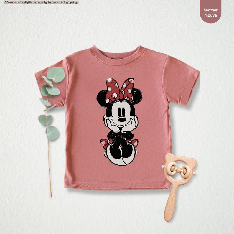 May include: Heather mauve t-shirt featuring a Minnie Mouse graphic. The graphic shows Minnie Mouse in black and white with a red polka dot bow. The shirt is short-sleeved and has a round neckline. A wooden rattle is next to the shirt.