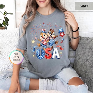 May include: Gray t-shirt featuring a patriotic design with two cartoon chipmunks celebrating with fireworks and the word "USA". The chipmunks are dressed in red, white, and blue outfits, holding flags and star-shaped balloons.