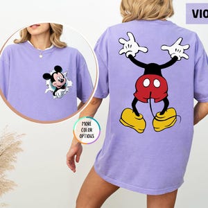 May include: A violet-coloured t-shirt with Mickey Mouse graphics. The front features Mickey breaking through a hole, and the back shows Mickey in a playful pose. The shirt has a comfortable, oversized fit.
