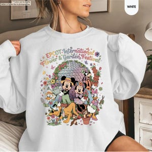 May include: White sweatshirt featuring a colorful graphic of Disney characters, including Mickey and Minnie Mouse, Goofy, and Donald Duck, surrounded by flowers and the text "EPCOT International Flower & Garden Festival."