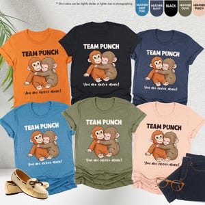 Puede incluir: Una colecci&oacute;n de camisetas en varios colores, cada una con un gr&aacute;fico de un mono de dibujos animados abrazando a otro mono. El texto "TEAM PUNCH" y "You are never alone!" est&aacute;n impresos encima y debajo del gr&aacute;fico. Un par de zapatos, unos vaqueros y unas gafas tambi&eacute;n est&aacute;n en la imagen.