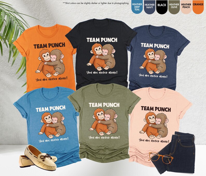Puede incluir: Una colecci&oacute;n de camisetas en varios colores, cada una con un gr&aacute;fico de un mono de dibujos animados abrazando a otro mono. El texto "TEAM PUNCH" y "You are never alone!" est&aacute;n impresos encima y debajo del gr&aacute;fico. Un par de zapatos, unos vaqueros y unas gafas tambi&eacute;n est&aacute;n en la imagen.