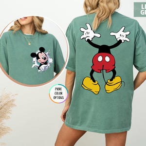 May include: Light green t-shirt with a Mickey Mouse design. The front has a small Mickey Mouse graphic, and the back features a larger image of Mickey Mouse with his arms raised. The shirt has a loose fit.