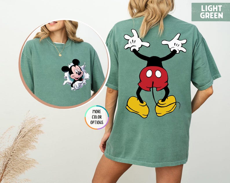 May include: Light green t-shirt with a Mickey Mouse design. The front has a small Mickey Mouse graphic, and the back features a larger image of Mickey Mouse with his arms raised. The shirt has a loose fit.