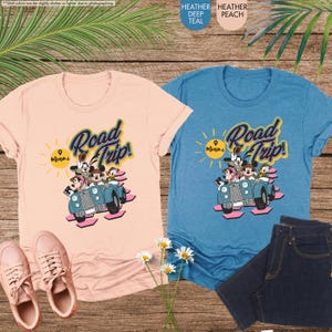 Op de afbeelding: Twee T-shirts, &eacute;&eacute;n perzik en &eacute;&eacute;n teal, tonen een afbeelding van stripfiguren in een vintage auto met de woorden "Road Trip! Miami." Het perzikkleurige shirt wordt gecombineerd met roze sneakers, terwijl het teal shirt naast een spijkerbroek staat.