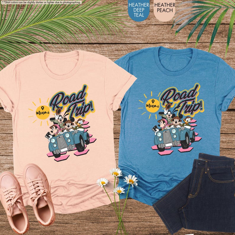 Op de afbeelding: Twee T-shirts, &eacute;&eacute;n perzik en &eacute;&eacute;n teal, tonen een afbeelding van stripfiguren in een vintage auto met de woorden "Road Trip! Miami." Het perzikkleurige shirt wordt gecombineerd met roze sneakers, terwijl het teal shirt naast een spijkerbroek staat.