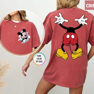 May include: Crimson-coloured t-shirt with a Mickey Mouse graphic. The front shows Mickey emerging from a torn hole, and the back displays a full-body cartoon image of Mickey Mouse. The shirt has short sleeves and a relaxed fit. The word 'CRIMSON' is in the top right corner.