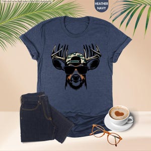 May include: A navy blue t-shirt with a graphic of a deer wearing sunglasses and a camouflage baseball cap. The deer has large antlers.