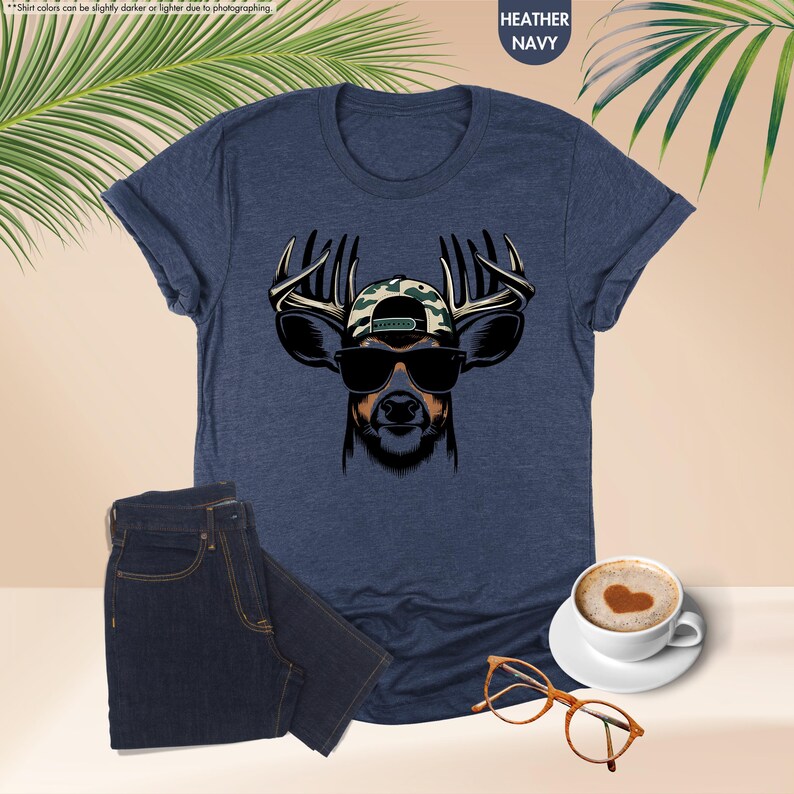 May include: A navy blue t-shirt with a graphic of a deer wearing sunglasses and a camouflage baseball cap. The deer has large antlers.