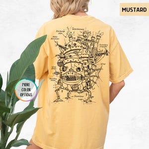 Op de afbeelding: Mosterdkleurig T-shirt met een gedetailleerde zwart-wit illustratie van een fantastisch bewegend huis, met labels zoals "Watchtower" en "Wings". Het shirt heeft ook de tekst "MORE COLOR OPTIONS" in een cirkel.