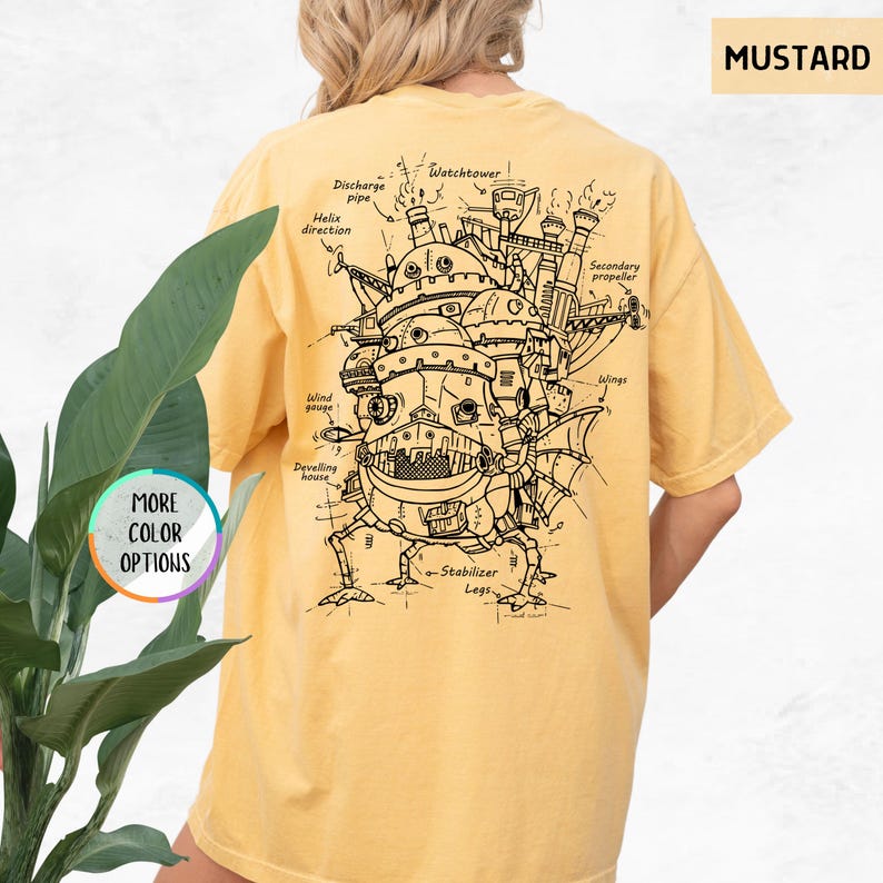 Op de afbeelding: Mosterdkleurig T-shirt met een gedetailleerde zwart-wit illustratie van een fantastisch bewegend huis, met labels zoals "Watchtower" en "Wings". Het shirt heeft ook de tekst "MORE COLOR OPTIONS" in een cirkel.