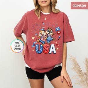 May include: Crimson-colored t-shirt featuring a patriotic design with the word "USA" in red, white, and blue. The graphic includes cartoon characters, fireworks, and stars, perfect for celebrating Independence Day.