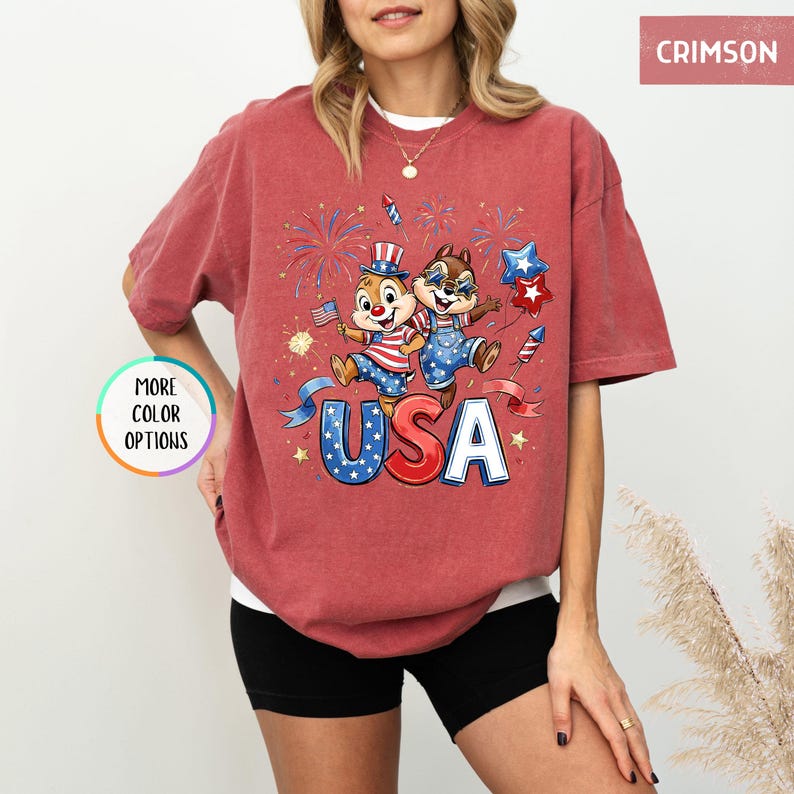 May include: Crimson-colored t-shirt featuring a patriotic design with the word "USA" in red, white, and blue. The graphic includes cartoon characters, fireworks, and stars, perfect for celebrating Independence Day.