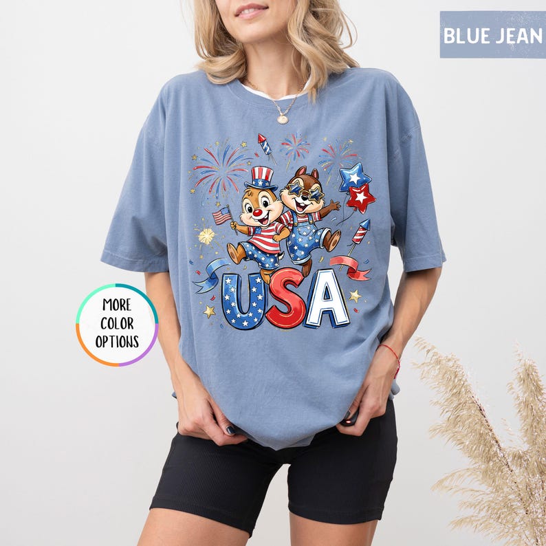 May include: Blue jean colored t-shirt with a graphic of two cartoon chipmunks celebrating with fireworks, flags, and the word "USA". The shirt has a round neck and short sleeves. The words "More Color Options" are in a circle on the left.