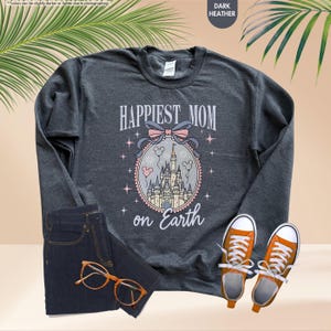 May include: Dark heather gray sweatshirt with the text "Happiest Mom on Earth" above a castle graphic. The sweatshirt is paired with dark blue jeans, orange sneakers, and glasses.