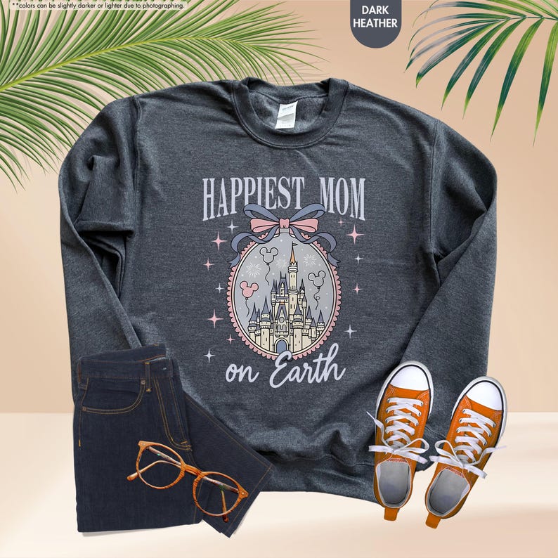 May include: Dark heather gray sweatshirt with the text "Happiest Mom on Earth" above a castle graphic. The sweatshirt is paired with dark blue jeans, orange sneakers, and glasses.