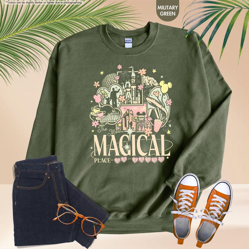 Pode incluir: Camisola verde militar com um gr&aacute;fico colorido que diz "The Most Magical Place". O design inclui um castelo, Mickey Mouse e outros elementos do parque tem&aacute;tico. Combinada com jeans azul escuro, t&ecirc;nis laranja e &oacute;culos.