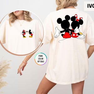 May include: Ivory-coloured t-shirt featuring Mickey and Minnie Mouse. The front displays a small graphic of the characters kissing, while the back showcases a larger image of them embracing. The shirt has a relaxed fit.