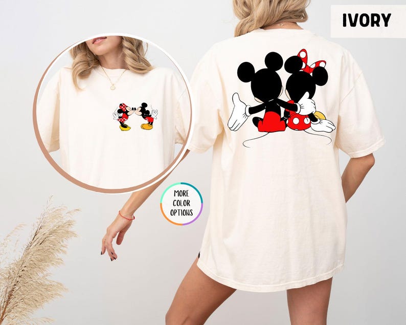 May include: Ivory-coloured t-shirt featuring Mickey and Minnie Mouse. The front displays a small graphic of the characters kissing, while the back showcases a larger image of them embracing. The shirt has a relaxed fit.
