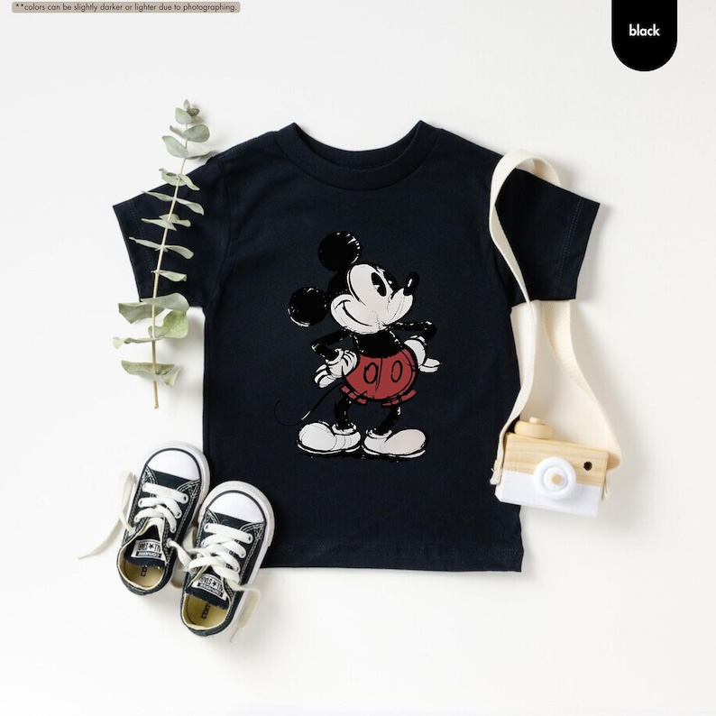 May include: Black toddler t-shirt featuring a classic Mickey Mouse graphic. The design includes Mickey's face, red shorts, and white gloves. The image also shows a pair of black and white sneakers and a small tote bag with a wooden camera.