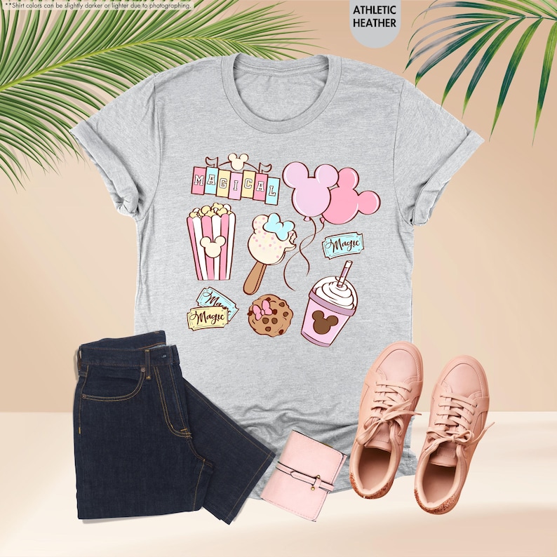 Puede incluir: Una camiseta gris jaspeada con un dise&ntilde;o de dibujos animados con Mickey Mouse y Minnie Mouse. El dise&ntilde;o incluye palomitas de ma&iacute;z, un helado de Mickey Mouse, globos y un batido de Mickey Mouse. El texto "Magical" aparece en un cartel del dise&ntilde;o.