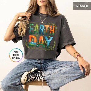 May include: A woman wearing a charcoal grey t-shirt with the text "Earth Day" in colorful letters with a nature scene design. The text is surrounded by green leaves. The woman is wearing blue jeans and black sneakers.