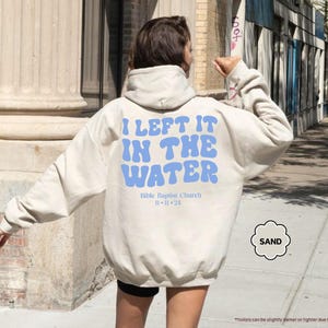 May include: Sand-colored hoodie with the text "I LEFT IT IN THE WATER" in blue, along with "Bible Baptist Church 11-11-24". The hoodie is a neutral color and has a drawstring hood.
