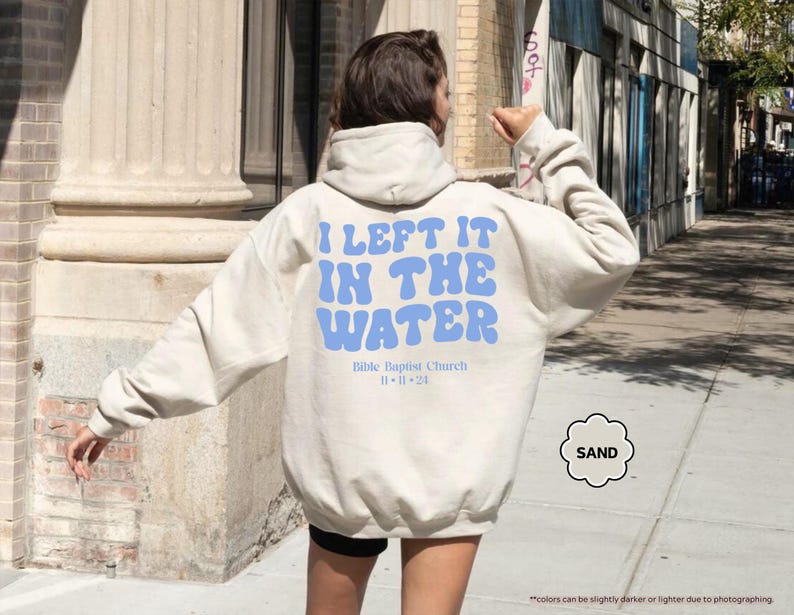 May include: Sand-colored hoodie with the text "I LEFT IT IN THE WATER" in blue, along with "Bible Baptist Church 11-11-24". The hoodie is a neutral color and has a drawstring hood.