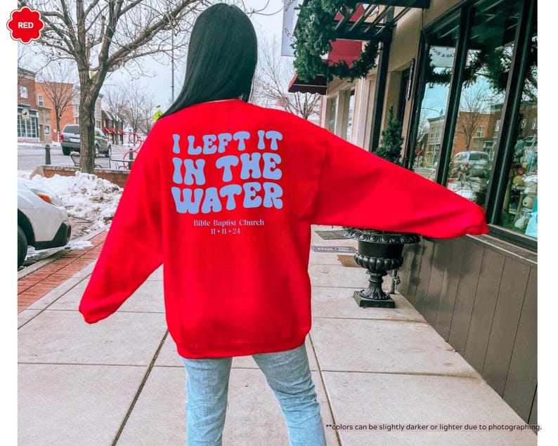 May include: A bright red sweatshirt with the text "I LEFT IT IN THE WATER" in light blue, along with "Bible Baptist Church 11-11-24". The sweatshirt is worn by a person standing outside.