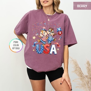 May include: Berry-colored t-shirt featuring a patriotic design with two cartoon chipmunks, fireworks, and the word "USA" in red, white, and blue. The shirt has a relaxed fit and short sleeves, perfect for casual wear.