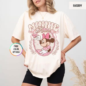 May include: A white t-shirt with a pink and brown graphic of Minnie Mouse holding a teacup. The graphic is surrounded by a pink floral frame and the text "Minnie Mouse" is above the graphic. The shirt is available in ivory.