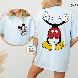 May include: Light blue t-shirt featuring Mickey Mouse. The front has a graphic of Mickey Mouse bursting through a hole. The back has a large graphic of Mickey Mouse with his arms up. The shirt is labelled "CHAMBRAY" and "MORE COLOR OPTIONS".