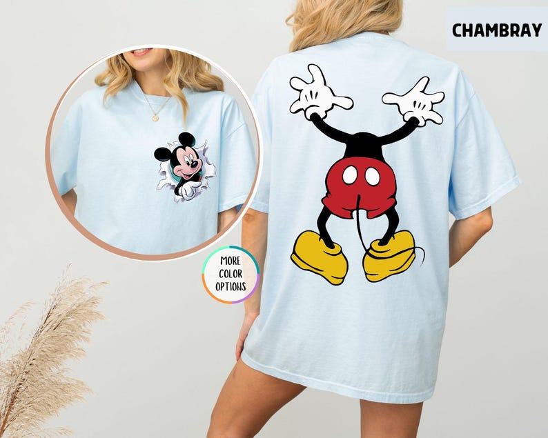May include: Light blue t-shirt featuring Mickey Mouse. The front has a graphic of Mickey Mouse bursting through a hole. The back has a large graphic of Mickey Mouse with his arms up. The shirt is labelled "CHAMBRAY" and "MORE COLOR OPTIONS".
