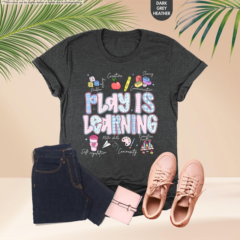 May include: Dark grey heather t-shirt with the text "PLAY IS LEARNING" in a colourful design. The image includes a pair of pink trainers, dark blue jeans, and a pink wallet. The shirt has illustrations of learning concepts.