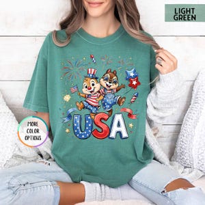 May include: Light green t-shirt featuring a graphic of two cartoon chipmunks celebrating with fireworks and the word "USA" in red, white, and blue. The shirt has a relaxed fit and short sleeves.
