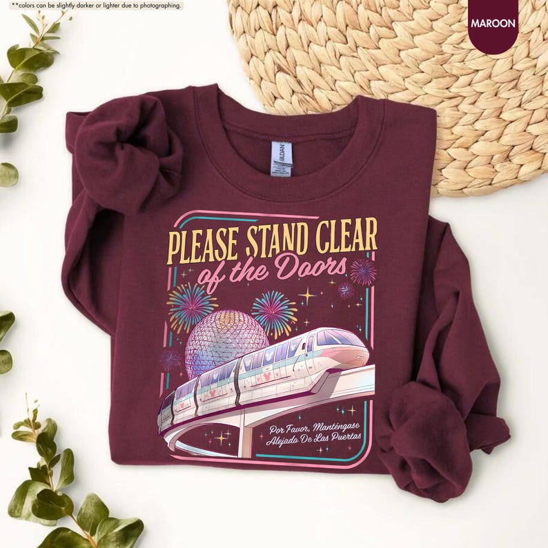 May include: Maroon sweatshirt with the text "PLEASE STAND CLEAR of the Doors" and a graphic of a monorail and fireworks. The sweatshirt also has the text "Por Favor. Mantingase Alejado De Las Puertas".