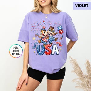 May include: Violet-colored t-shirt featuring a patriotic graphic with two cartoon chipmunks, fireworks, and the word "USA" in red, white, and blue. The shirt has a relaxed fit and short sleeves. The image also includes a "More Color Options" button.