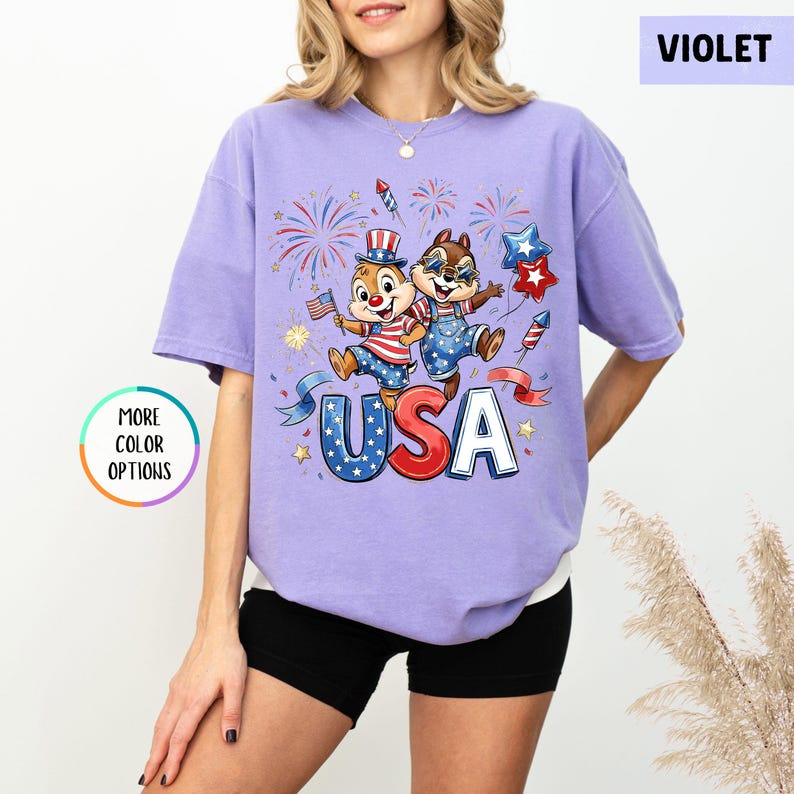 May include: Violet-colored t-shirt featuring a patriotic graphic with two cartoon chipmunks, fireworks, and the word "USA" in red, white, and blue. The shirt has a relaxed fit and short sleeves. The image also includes a "More Color Options" button.