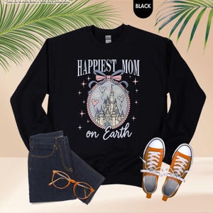 May include: Black sweatshirt with the text "Happiest Mom on Earth" in white script. The design includes a castle illustration with a pink bow and stars. The sweatshirt is paired with blue jeans and orange sneakers.