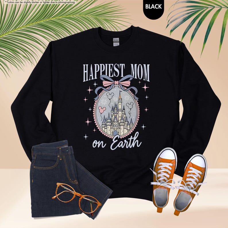 May include: Black sweatshirt with the text "Happiest Mom on Earth" in white script. The design includes a castle illustration with a pink bow and stars. The sweatshirt is paired with blue jeans and orange sneakers.