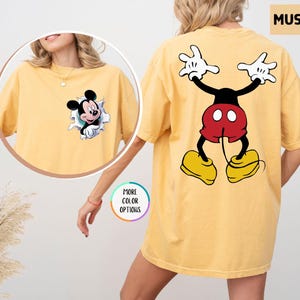 May include: Mustard-coloured t-shirt featuring Mickey Mouse. The front has a graphic of Mickey bursting through a torn paper effect. The back displays a large Mickey Mouse graphic in a playful pose. The shirt is a comfortable, oversized fit.