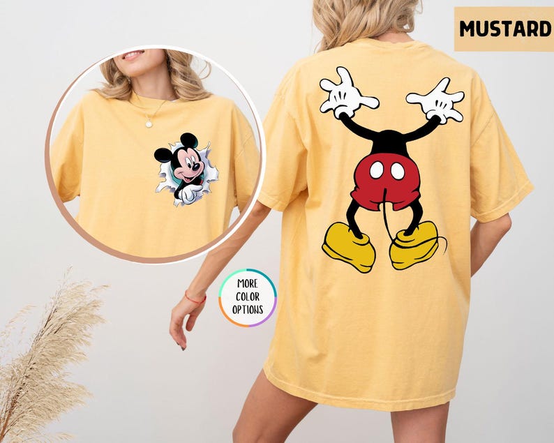 May include: Mustard-coloured t-shirt featuring Mickey Mouse. The front has a graphic of Mickey bursting through a torn paper effect. The back displays a large Mickey Mouse graphic in a playful pose. The shirt is a comfortable, oversized fit.
