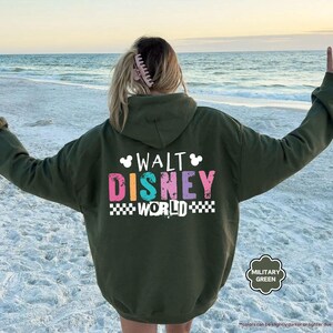Puede incluir: Sudadera con capucha verde militar con el texto "WALT DISNEY WORLD" en letras de bloque coloridas. La sudadera tiene una capucha con cord&oacute;n y un bolsillo de canguro. El fondo es una playa con el oc&eacute;ano.